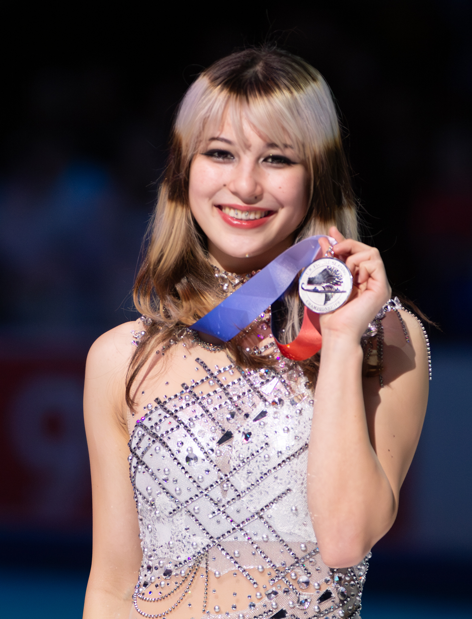 A figure skater smiles while holding up a medal on a red, white, and blue ribbon at the 2026 United States Figure Skating Championships. She wears a crystal-encrusted skating dress and has long light brown hair with bangs.