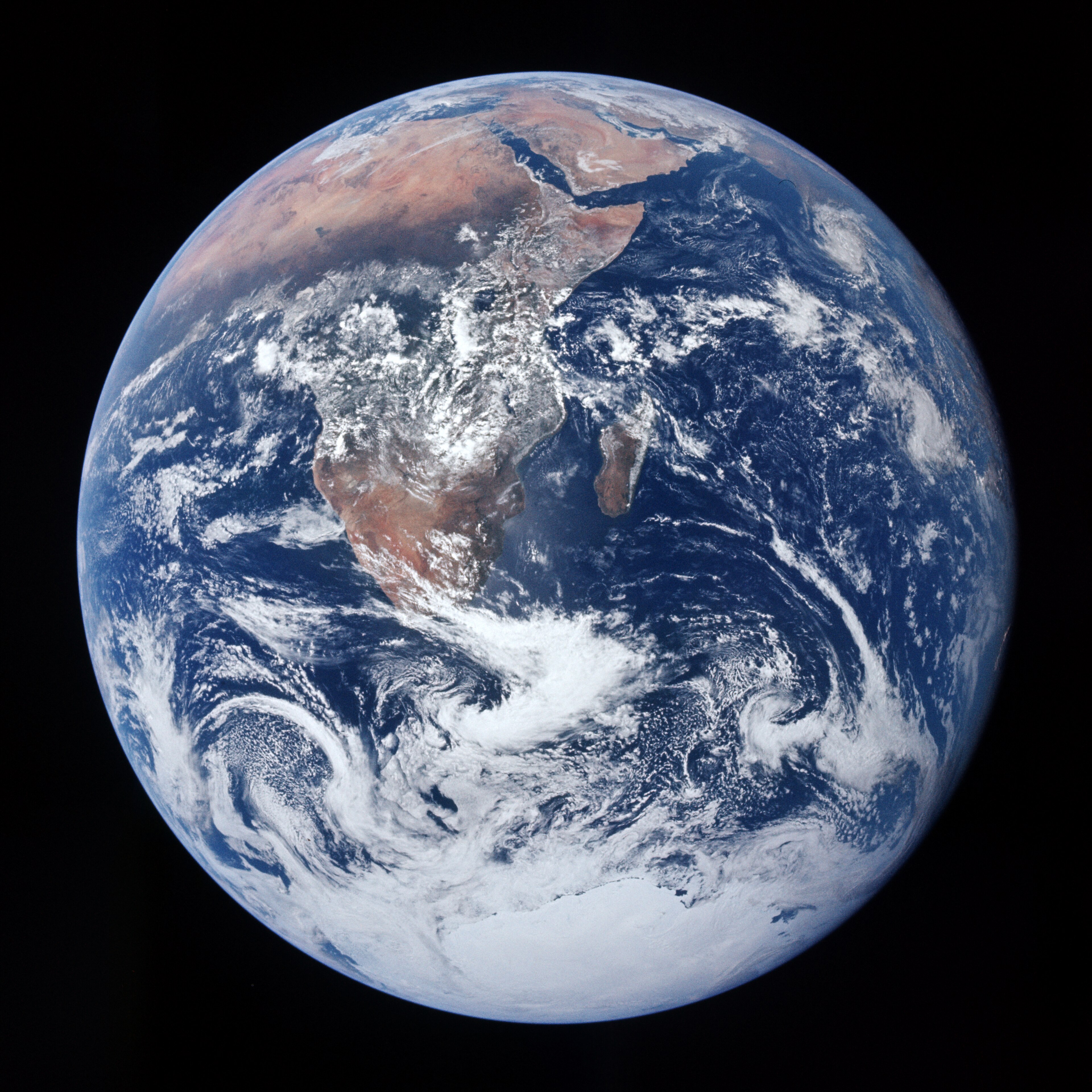 A full-disk photograph of Earth taken from space against a black background, showing the African continent prominently in the centre-right, with the Arabian Peninsula visible at the top. Swirling white cloud formations dominate the lower half, and Antarctica appears as a white mass at the bottom edge. The deep blue of the Indian and Atlantic Oceans contrasts with the reddish-brown of southern Africa's interior.