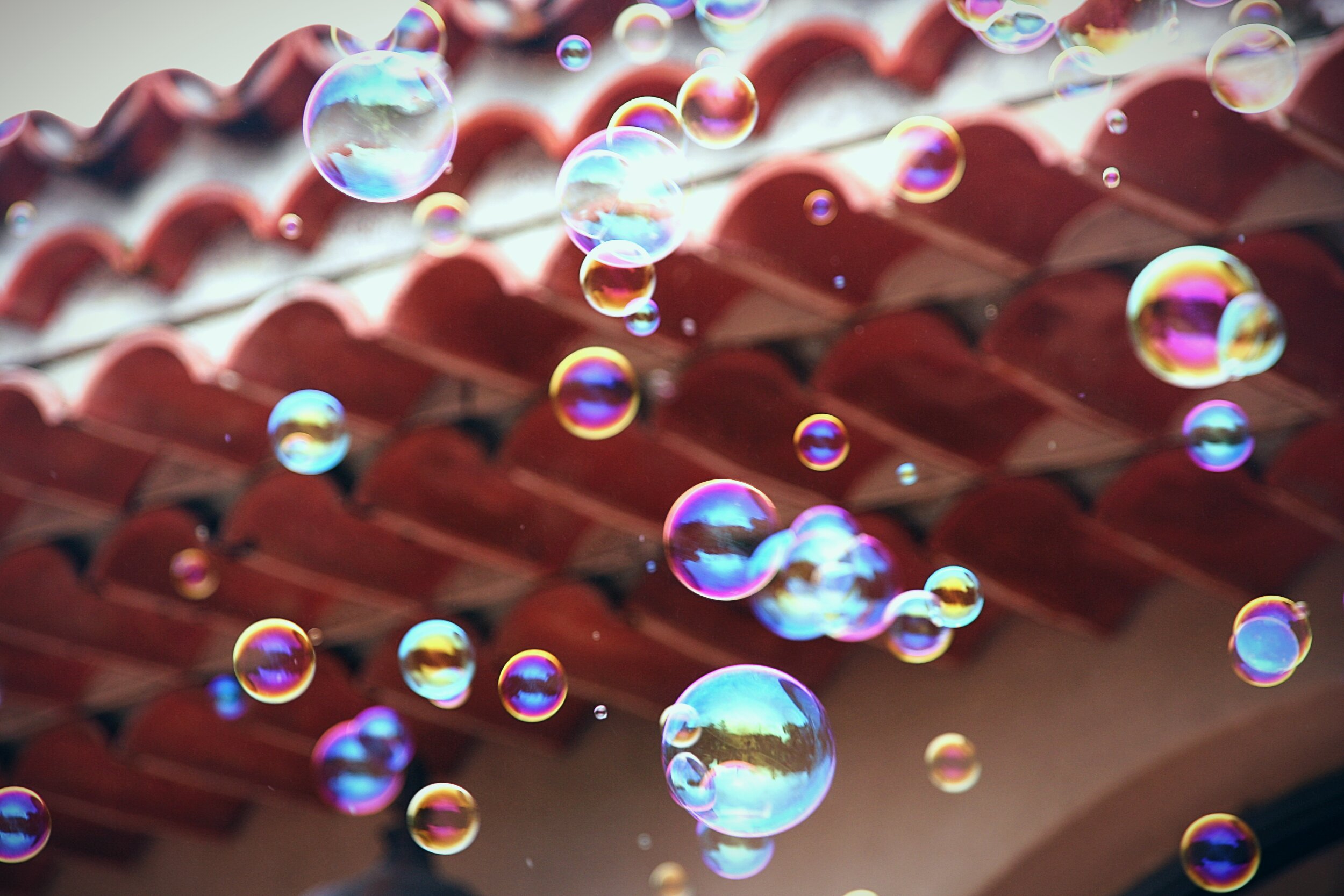 Dozens of soap bubbles float upward against a backdrop of terracotta roof tiles shot from below. The bubbles range in size from small to large and display iridescent colours: electric blue, magenta, gold, and green. All shimmering across their thin, transparent surfaces. Some bubbles reflect distorted glimpses of sky and surroundings. The roof tiles are rendered soft and out of focus in warm red-brown tones, creating contrast with the bright, jewel-like spheres in the foreground.