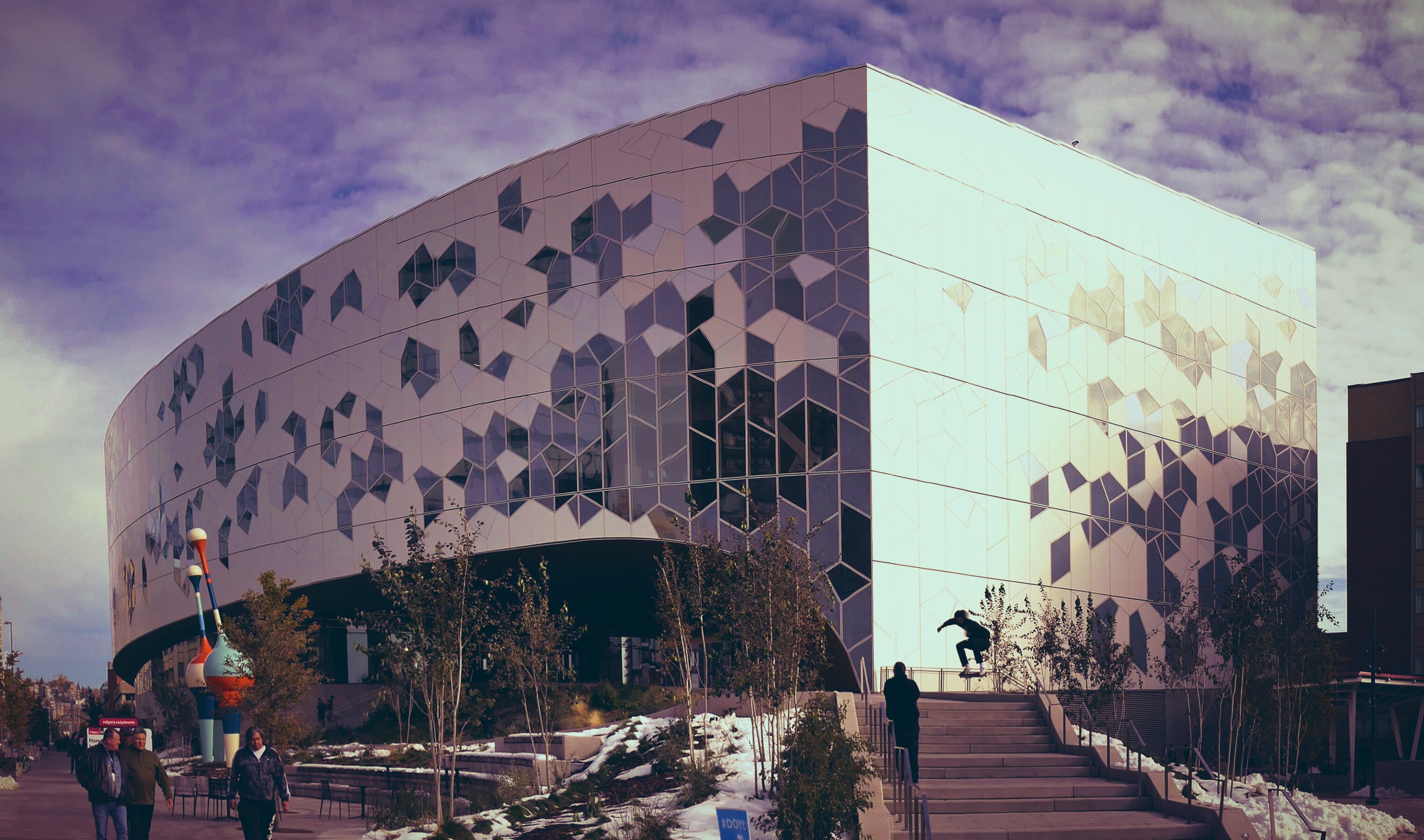 The Calgary Central Library, a striking curved building with a geometric glass and aluminum façade, on a partly cloudy autumn day. A skateboarder performs a trick on the front steps while pedestrians walk along the sidewalk below. Patches of snow are visible on the ground, and a colourful sculpture stands to the left of the entrance.