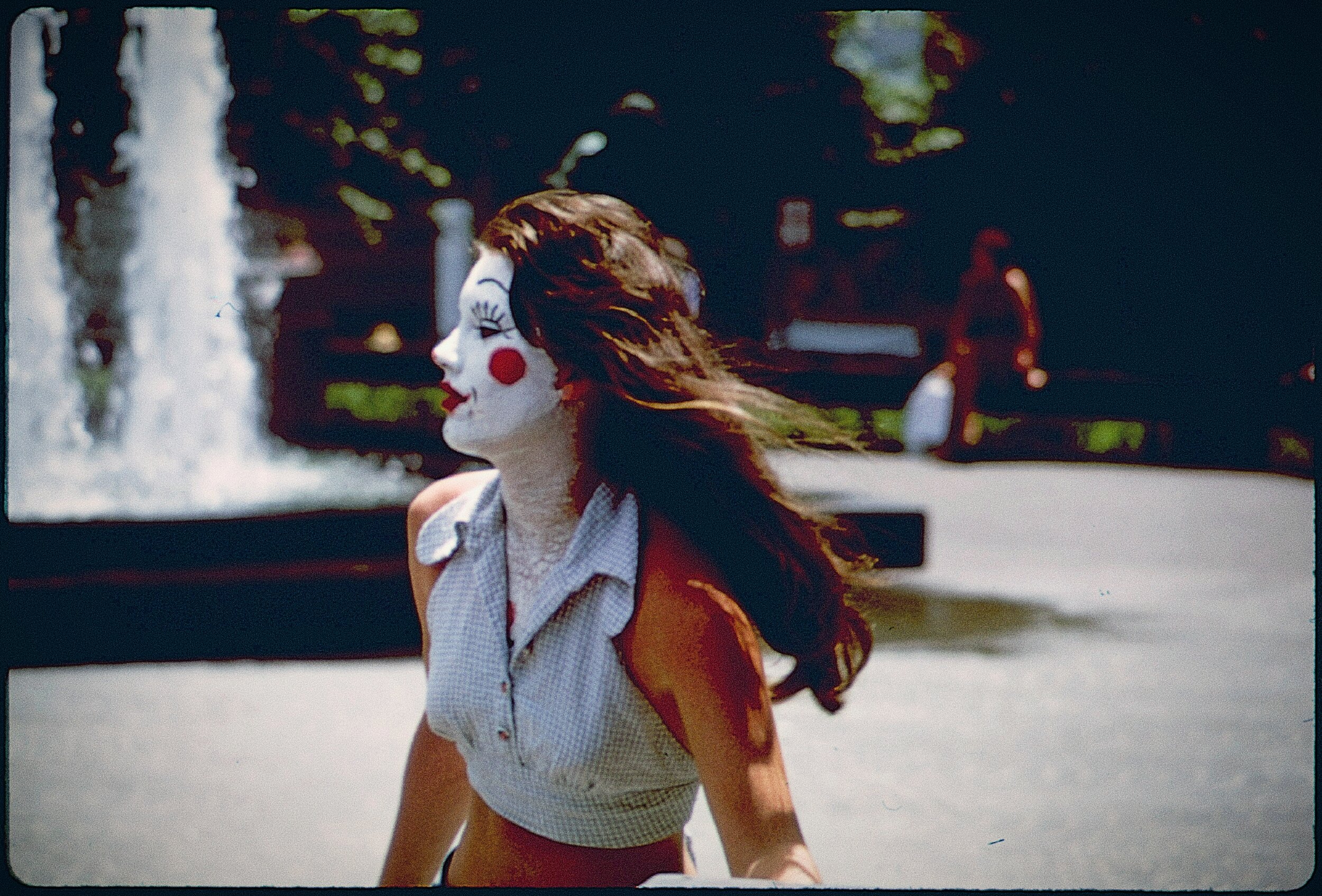 Vintage-toned photo of a young woman in profile with clown-style face paint, a red circle on her cheek and a painted winking eye. Her dark hair blowing sideways in the wind. She wears a light gingham sleeveless top and stands before a plaza fountain, with a blurred figure sitting in the background.