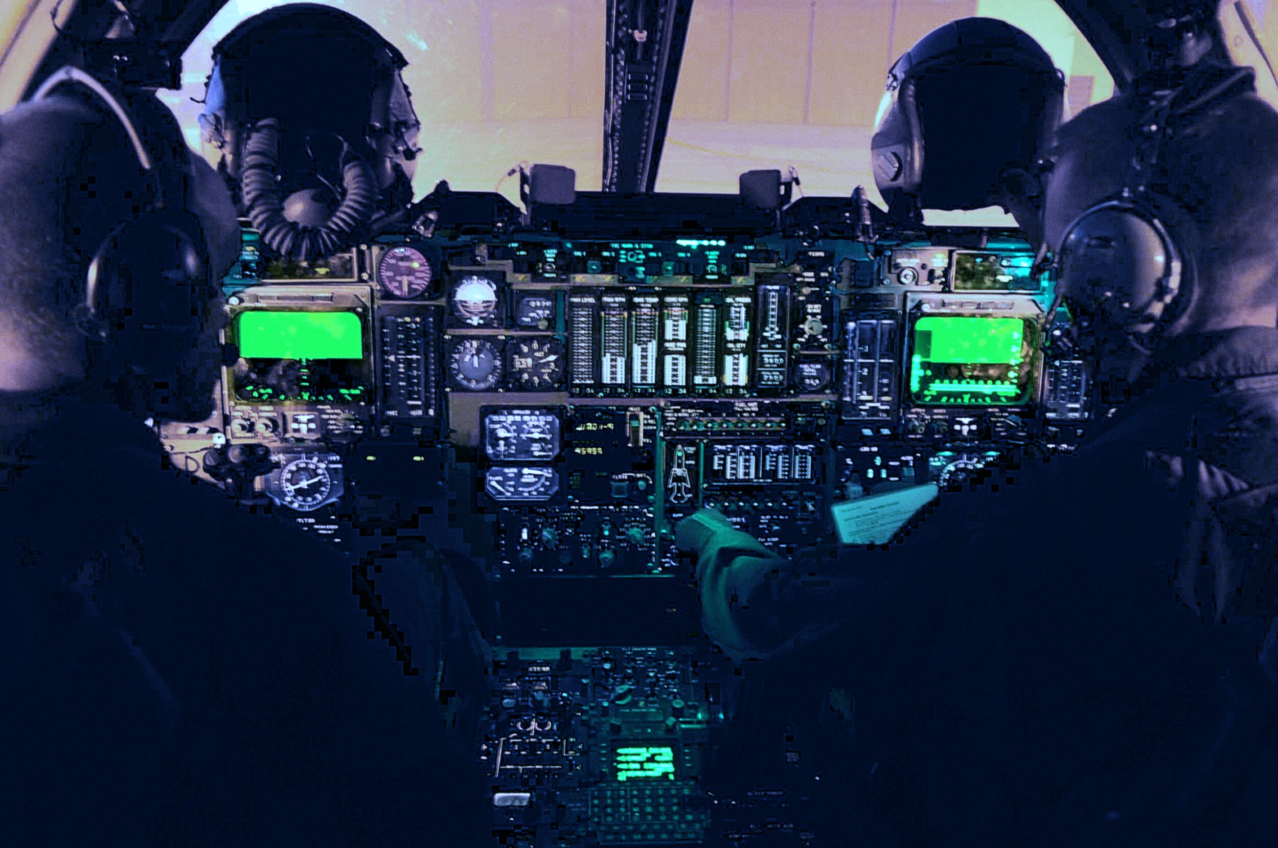 Two aircrew members wearing helmets, headsets, and oxygen masks are seated in a dimly lit military aircraft cockpit. The instrument panel between them glows with green multi-function displays, rows of analog engine gauges, and backlit switches, while light is visible through the windscreen ahead.