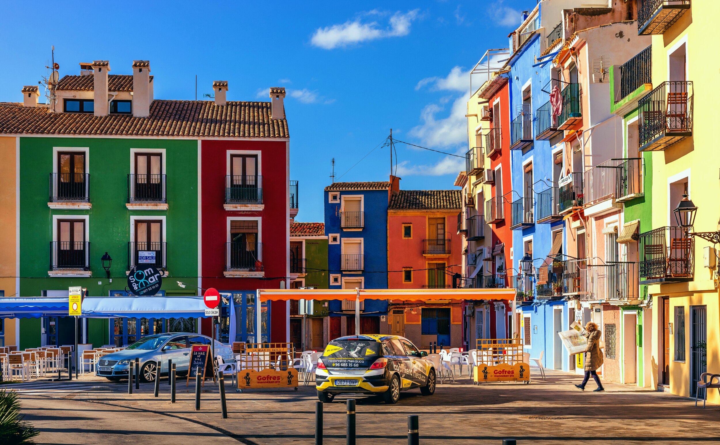 A sunny street in a Mediterranean fishing village lined with brightly painted row houses in green, red, blue, orange, and yellow, with wrought-iron balconies. Cafe tables and umbrellas sit along the sidewalk, two cars are parked on the cobblestone street, and a person in a coat walks past while reading a newspaper.