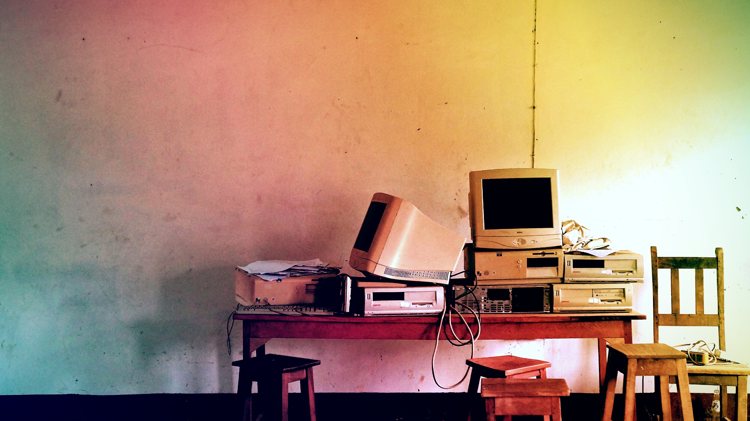 A cluttered wooden table holds a row of obsolete beige desktop computers and CRT monitors, including a Dell and Compaq, with tangled cables, stacked against a stained wall lit in a pink-to-yellow-to-green gradient. Wooden stools and a chair sit in front of the table, suggesting an abandoned classroom or office.