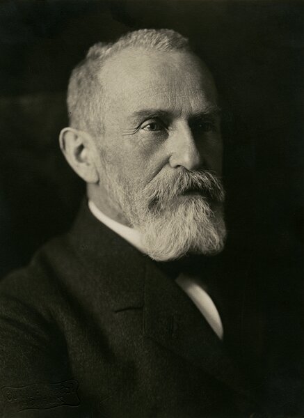 Formal black-and-white portrait of an older man with a receding hairline and a full white beard and mustache. He wears a dark suit and looks slightly to the side with a serious expression against a dark studio background.