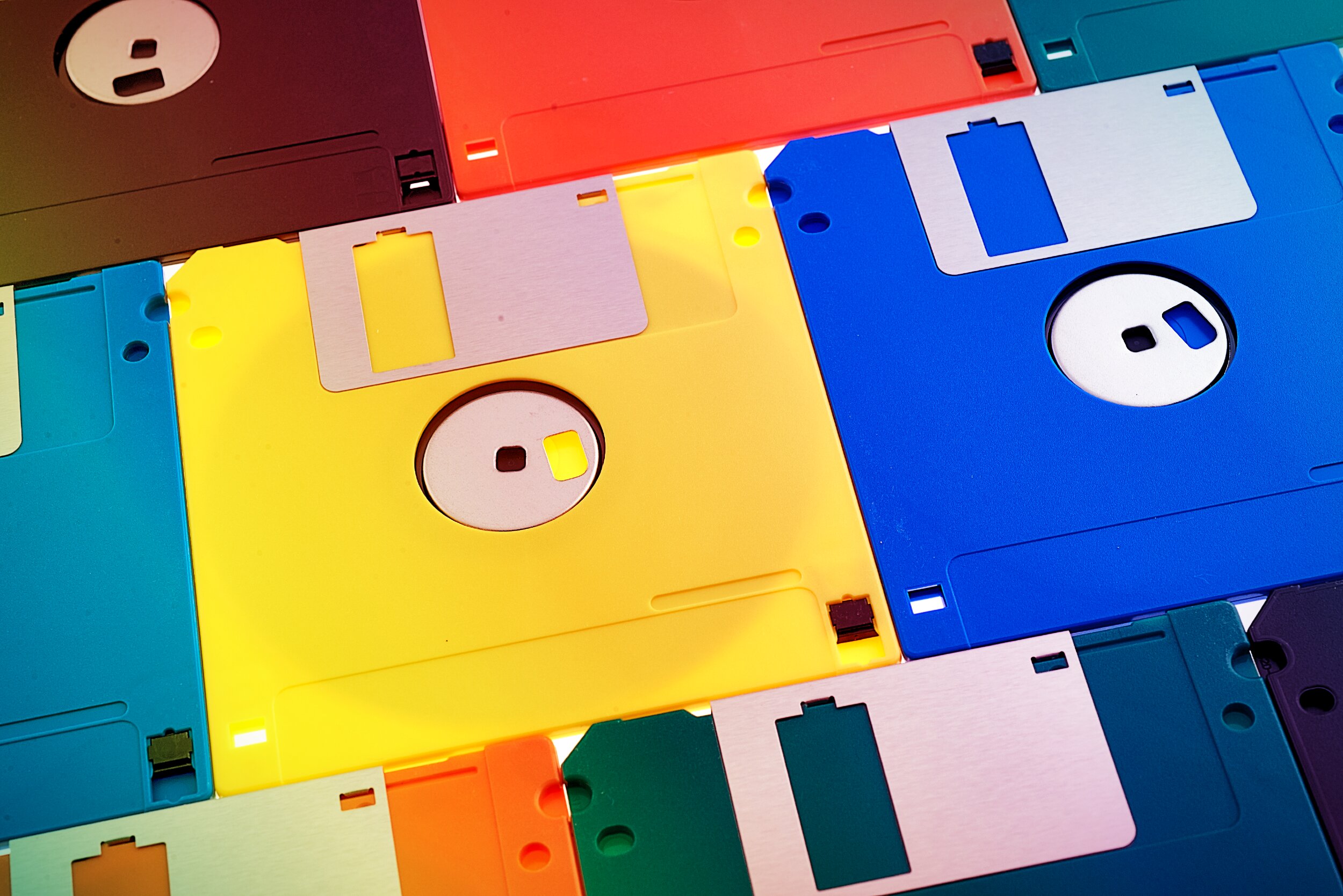 A collection of 3.5-inch floppy disks arranged in an overlapping, flat-lay pattern filling the entire frame. The disks span a wide range of colours — yellow, blue, red, teal, orange, dark brown, purple, and grey — creating a vibrant, saturated composition. The metal write-protect shutters and circular hub windows are visible on several disks, shot in close-up with bright, even lighting.