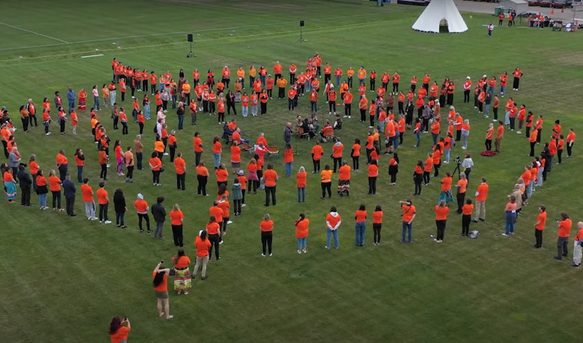 An aerial drone photo of a green sports field where many people, almost all in orange shirts, stand spaced apart in loose concentric rings that form the shape of a giant spiderweb. At the centre, a small group sits in a circle with drums and chairs. Some people wear ribbon skirts, someone on the right stands behind a camera on a tripod, and in the bottom-left foreground a woman in orange holds up her phone to photograph the web.