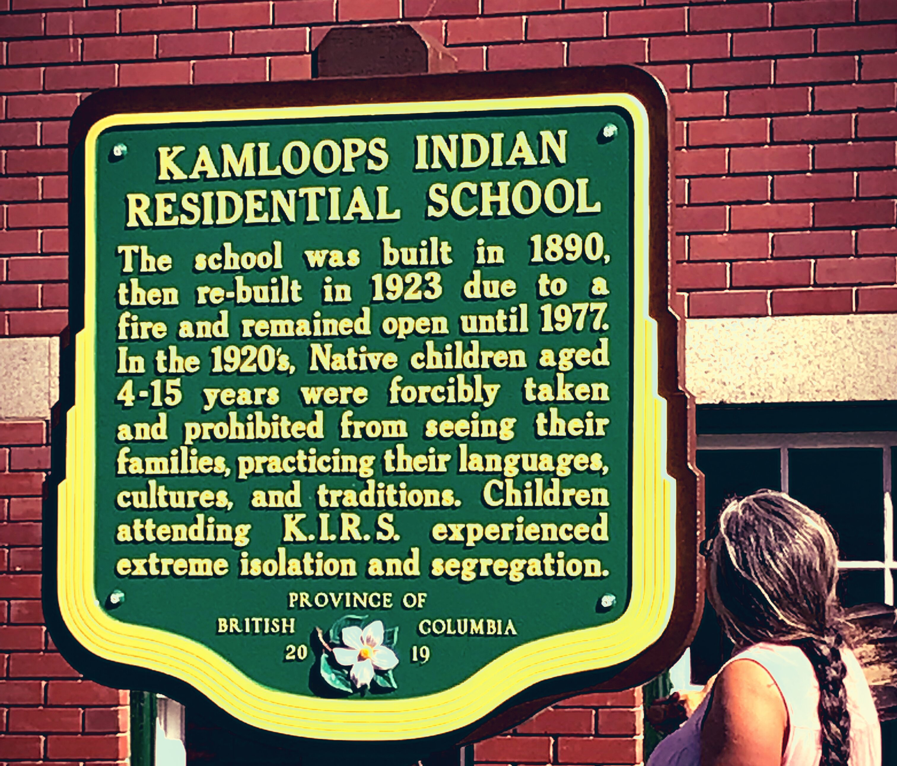 A green historical plaque mounted on a brick wall titled 'Kamloops Indian Residential School,' describing the school's operation from 1890 to 1977 and the forced removal, isolation, and cultural suppression of Indigenous children; a person stands partially visible to the right, looking at the sign