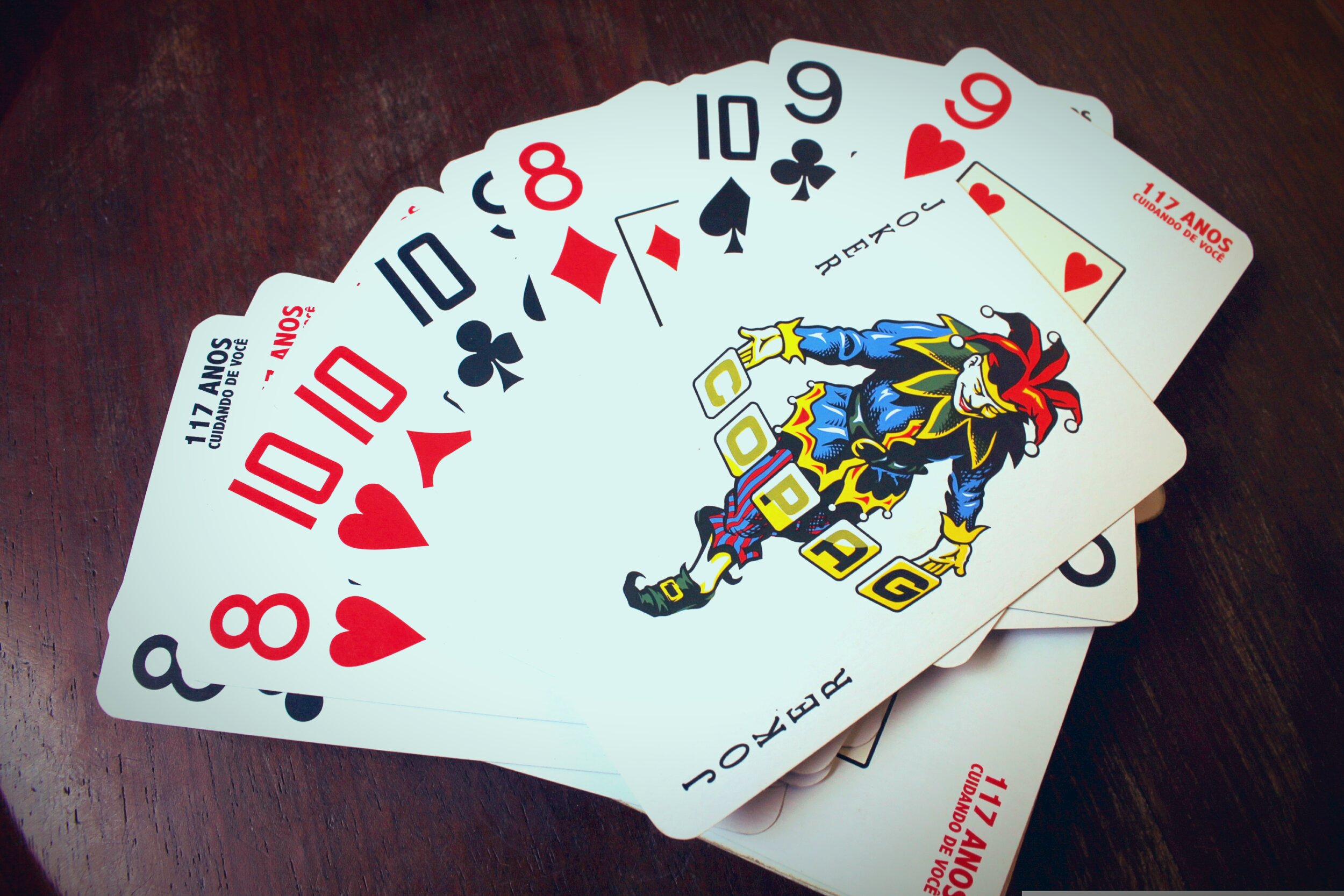 Several playing cards fanned out face-up on a dark wood surface. Visible cards from left to right include the 8 of Hearts, 10 of Hearts, 10 of Clubs, 8 of Diamonds, 10 of Spades, 9 of Clubs, and 9 of Hearts. Prominently displayed near the center is a Joker card depicting a jester figure in a colorful blue, green, and gold costume with a red and black jester's cap, holding letter blocks. Additional Joker and number cards are partially visible beneath the spread. All cards bear the text '117 Anos — Cuidando de Você,' a branded Brazilian playing card deck.