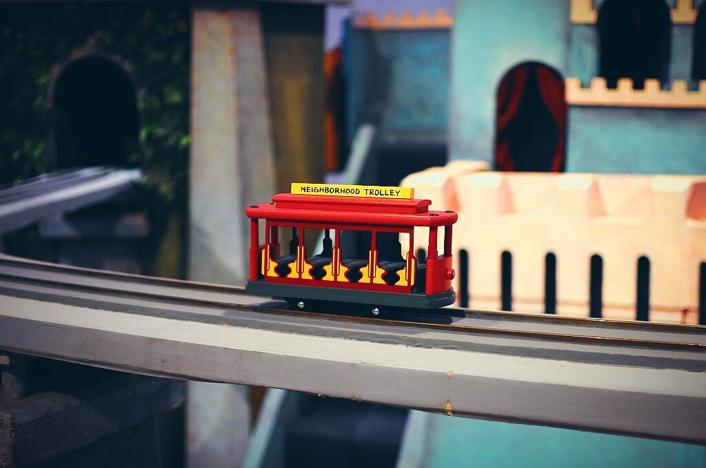 A red miniature trolley with 'NEIGHBORHOOD TROLLEY' written on a yellow sign on top, sitting on silver tracks. The trolley features yellow and black trim, visible passenger silhouettes in the windows, and is photographed against a blurred background of a miniature neighborhood set with colorful buildings.Shot taken during the 'Neighborhood of Make-Believe Tour' at WQED studios in Pittsburgh.