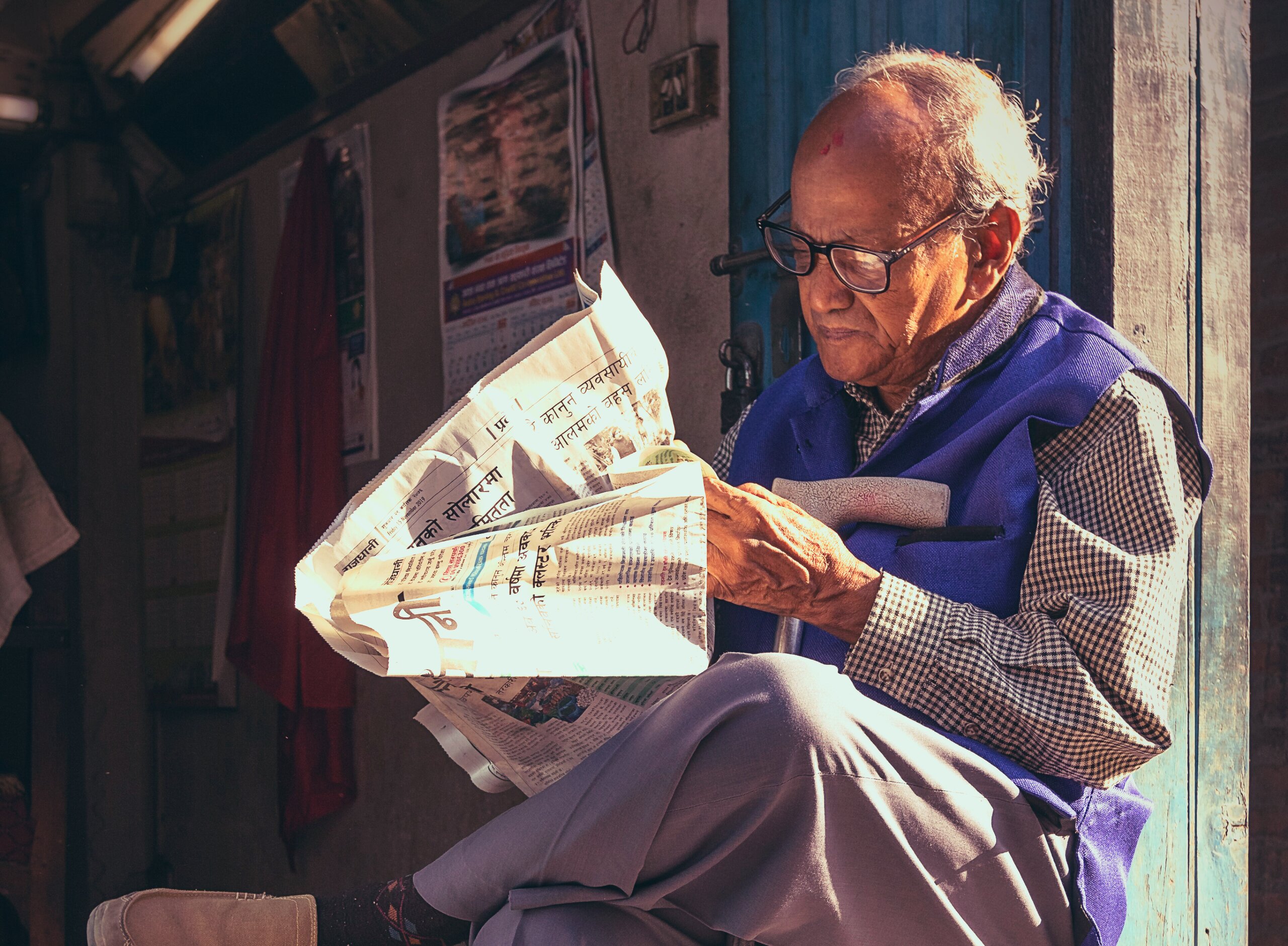 An elderly man sits in morning sunlight outside a weathered blue doorway in Basantapur, Nepal, absorbed in reading a Nepali-language newspaper. He wears black-rimmed glasses, a blue vest over a checked shirt, and light grey trousers. The newspaper catches the early light brilliantly against the dark background. A cane rests under his arm.