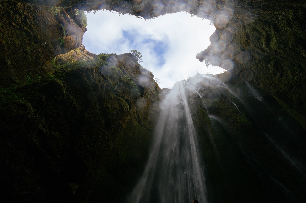 A dark pit with a waterfall cascading down steep moss-covered cliff walls, viewed from below, with mist rising through the narrow canyon opening to a cloudy sky above
