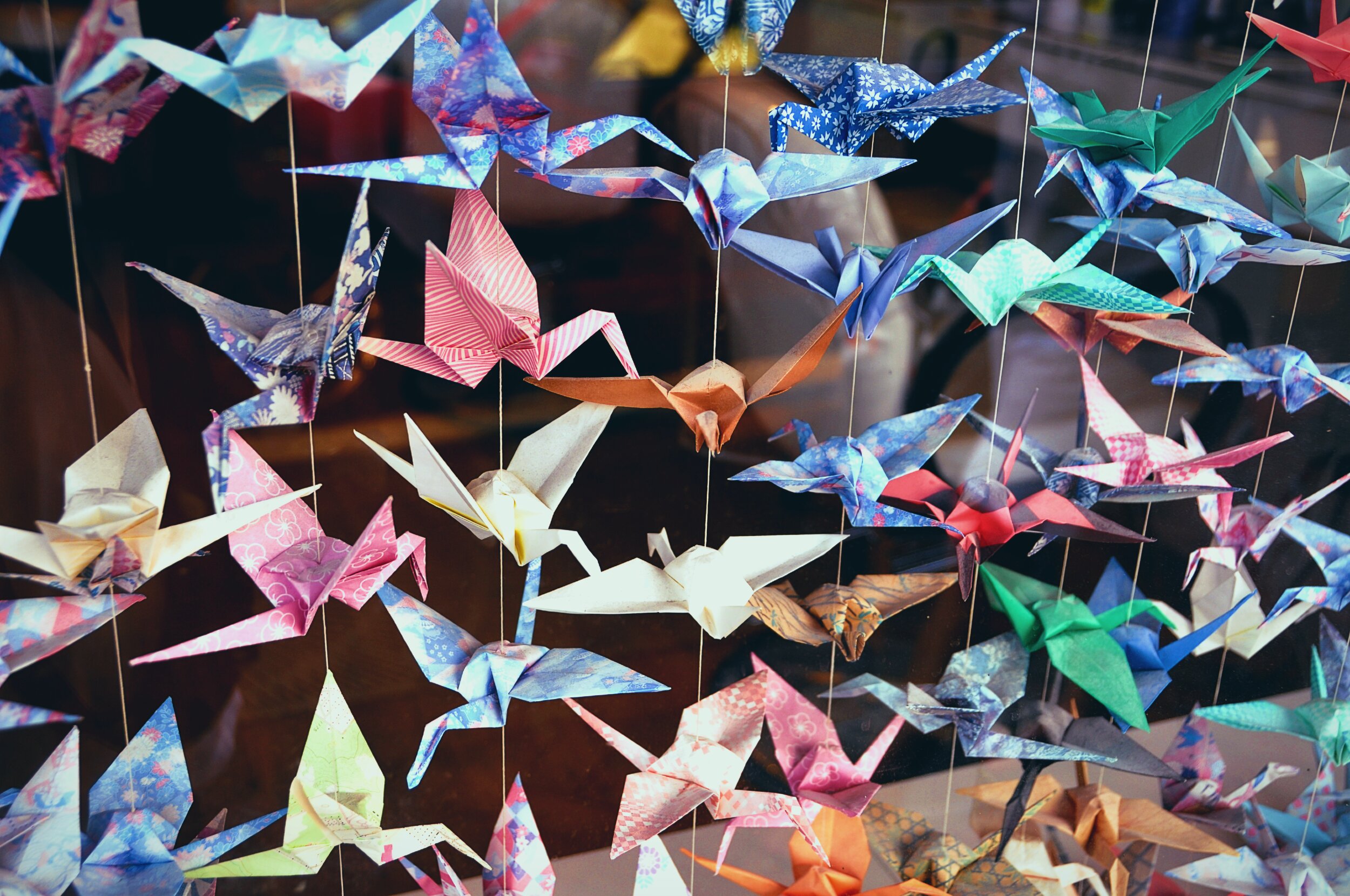 Dozens of origami paper cranes in a wide variety of colours and patterns. Blues, pinks, reds, greens, white, and earth tones, many made from patterned washi-style paper with florals and stripes, suspended from thin strings against a blurred warm-toned background.