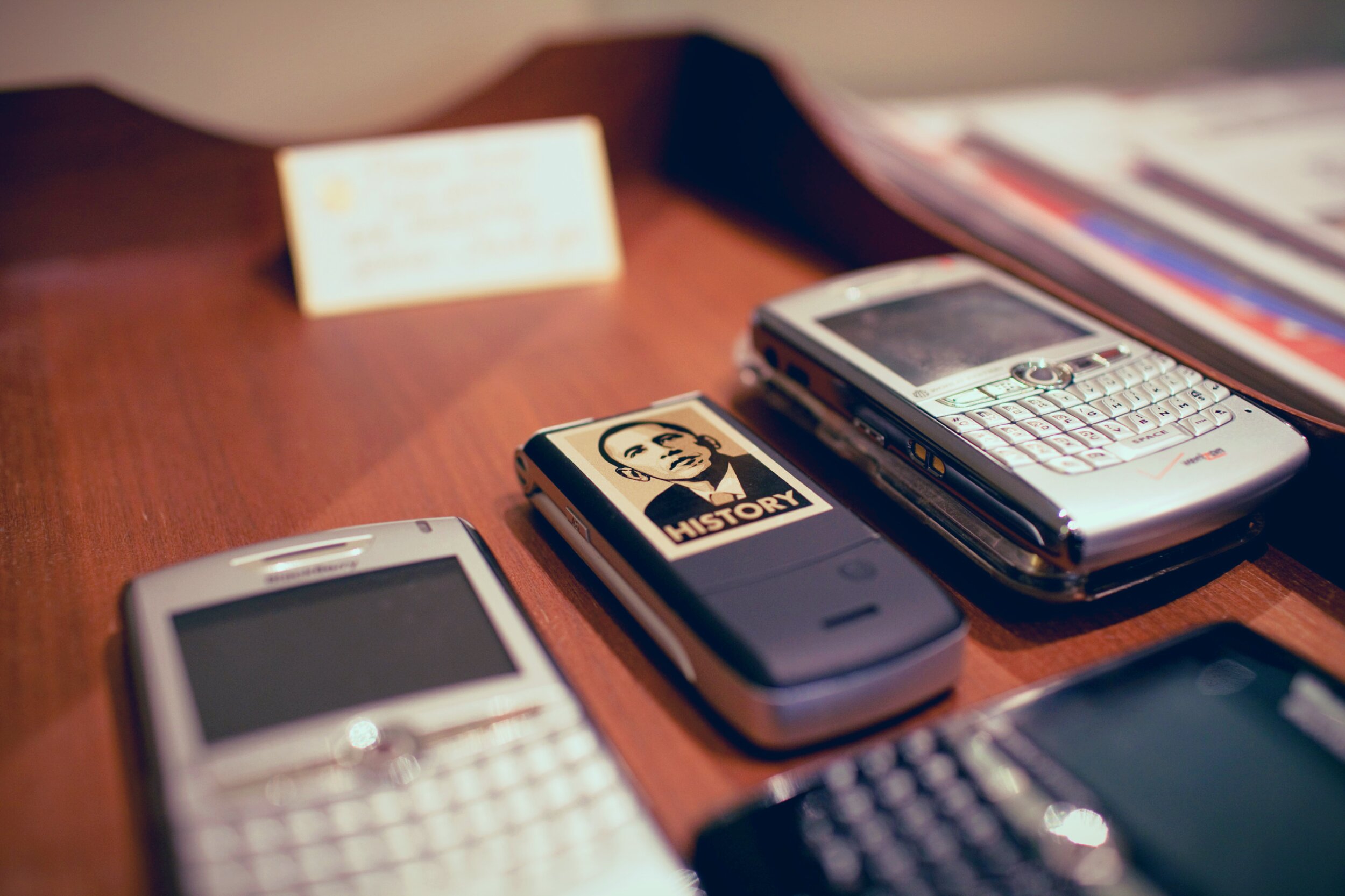 Four smartphones/PDAs from the mid-to-late 2000s arranged on a wooden desk. Two silver BlackBerry-style devices with physical QWERTY keyboards are open at top right and bottom left. A dark purple flip phone in the center has a sticker in the Shepard Fairey 'HOPE' poster style with the word 'HISTORY' beneath the portrait. A fourth device with a black keyboard is visible at bottom right. A blurred index card and papers sit in the background.