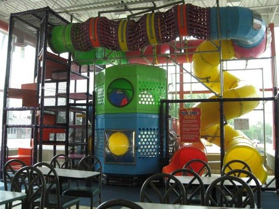 Indoor McDonald's PlayPlace structure, featuring a large multi-level climbing apparatus with colourful plastic tubes, tunnels, and a yellow spiral slide. The play structure is enclosed in a black metal frame and sits beside restaurant seating.
