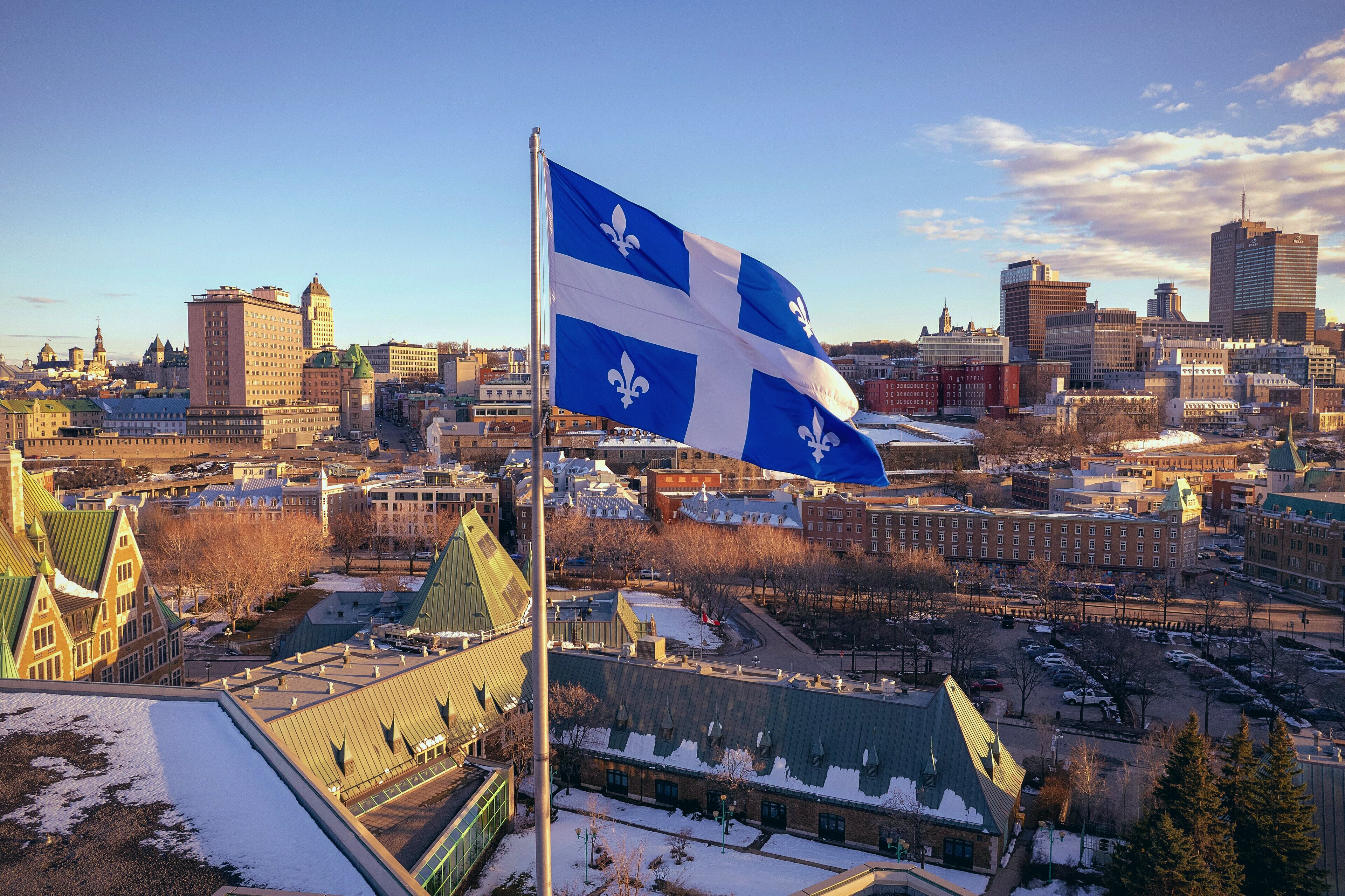 Le drapeau du Québec, le Fleurdelisé, bleu et blanc orné de quatre fleurs de lys, flotte au vent sur un mât au premier plan. En arrière-plan s'étend une vue aérienne de la ville de Québec en hiver : toits enneigés, architecture patrimoniale en briques rouges, toitures en cuivre vert aux pignons caractéristiques du Vieux-Québec, et la silhouette du centre-ville moderne à l'horizon, sous un ciel bleu parsemé de nuages dorés par la lumière rasante.