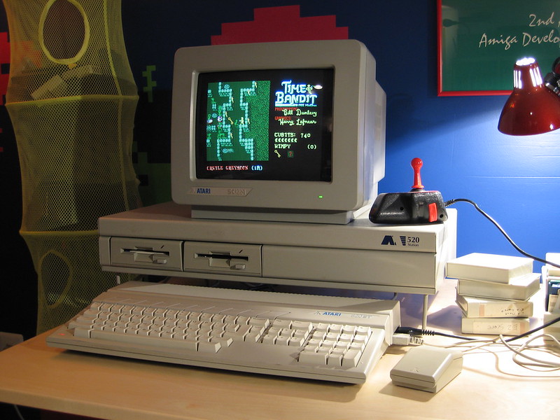Vintage Atari 520ST computer setup displaying Time Bandit game on CRT monitor, with keyboard, joystick with red button, and mouse on a desk. Yellow speaker visible on left and red desk lamp on right in background.