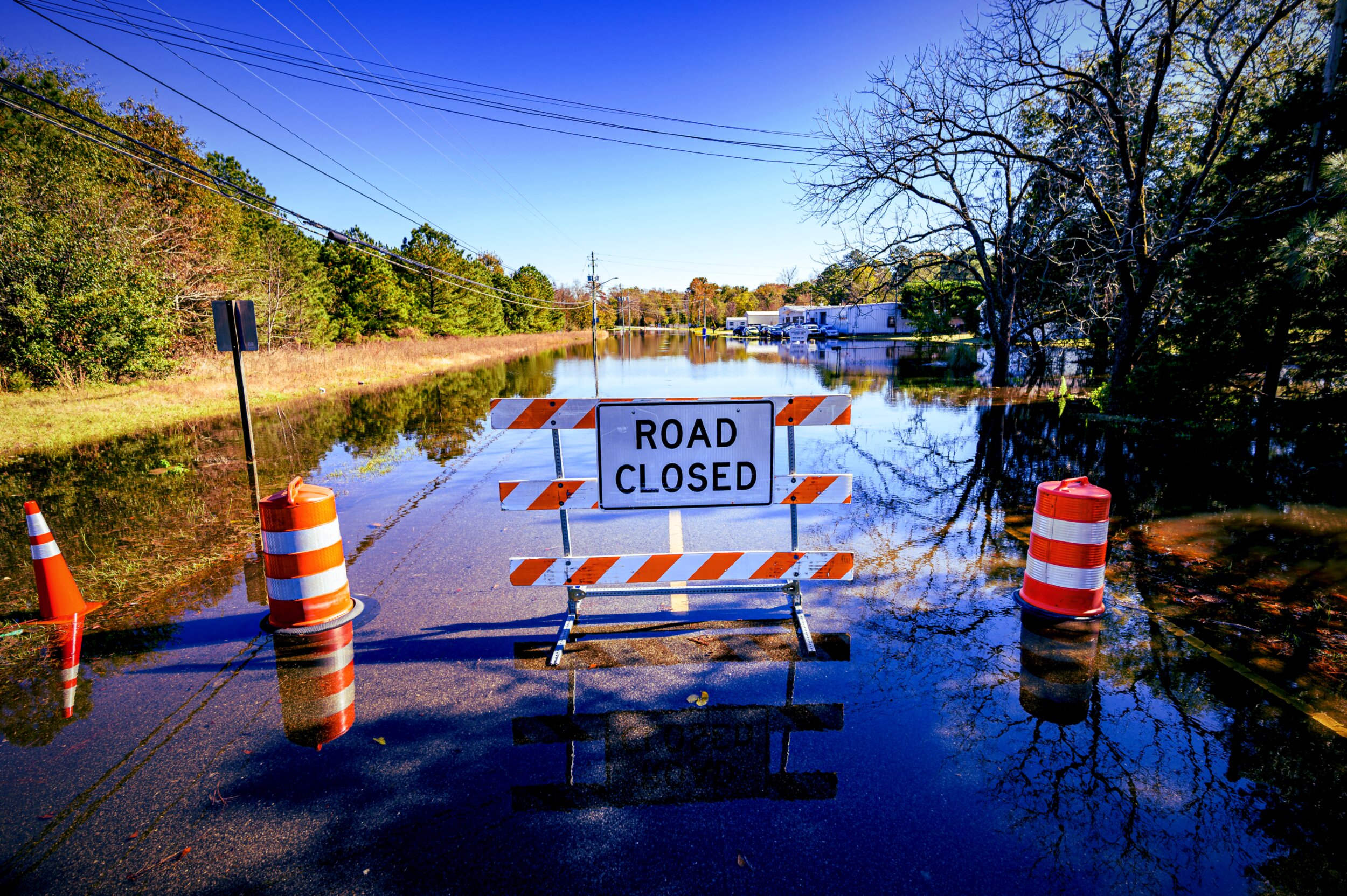A flooded roadway leading to a 'Road Closed' sign flanked by orange traffic barrels, with floodwater stretching to submerged buildings and parked cars in the distance under a clear blue sky.
