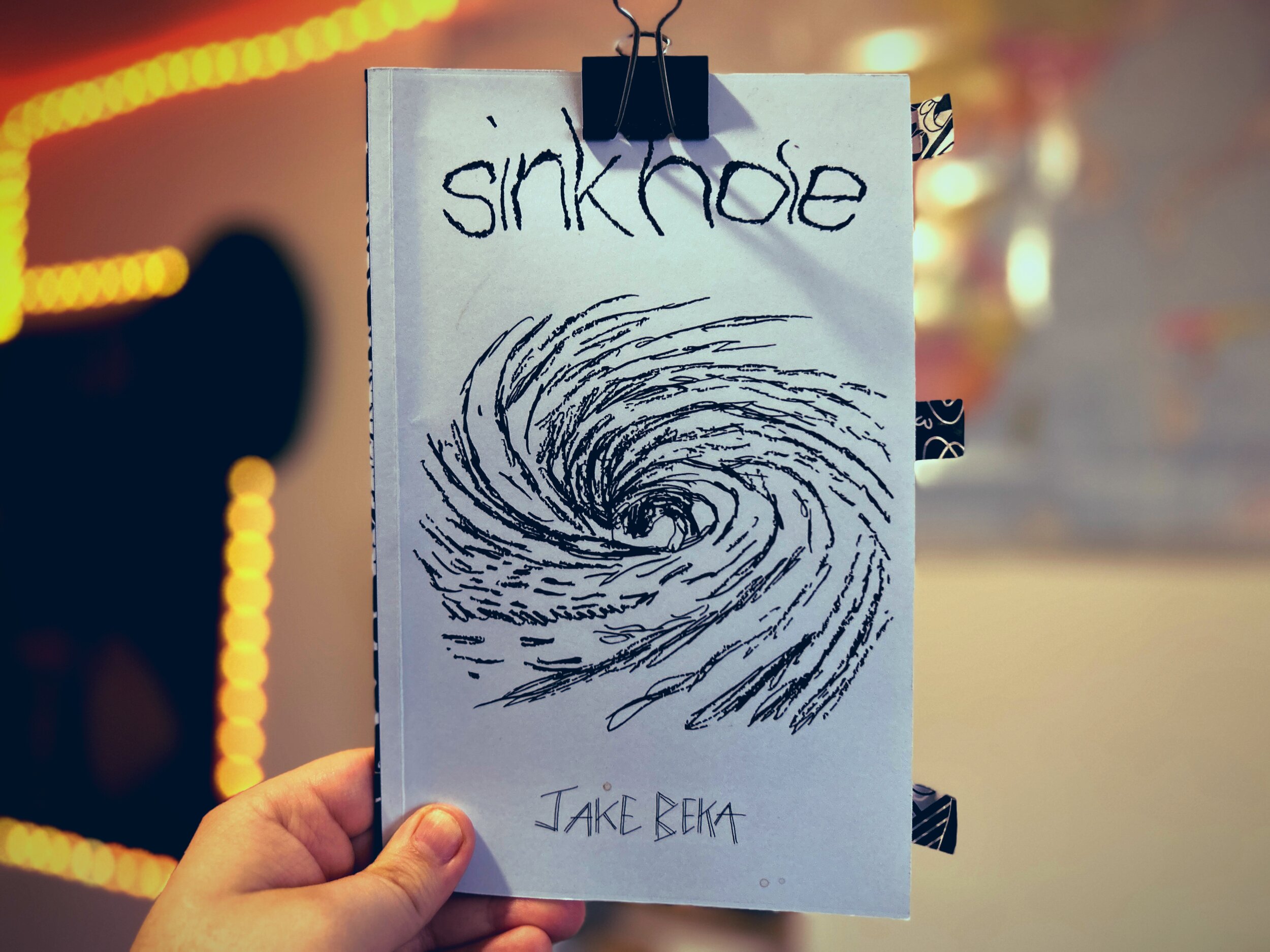 Hand holding a poetry chapbook titled “sinkhole,” the cover clipped at the top with a black binder clip. The light blue cover shows a loose black-ink drawing of a swirling vortex or whirlpool, with the title written in rough, hand-lettered lowercase at the top and “Jake Beka” handwritten at the bottom. Patterned tape tabs stick out from the right edge of the booklet. The background is softly blurred with warm yellow string lights forming curved lines.