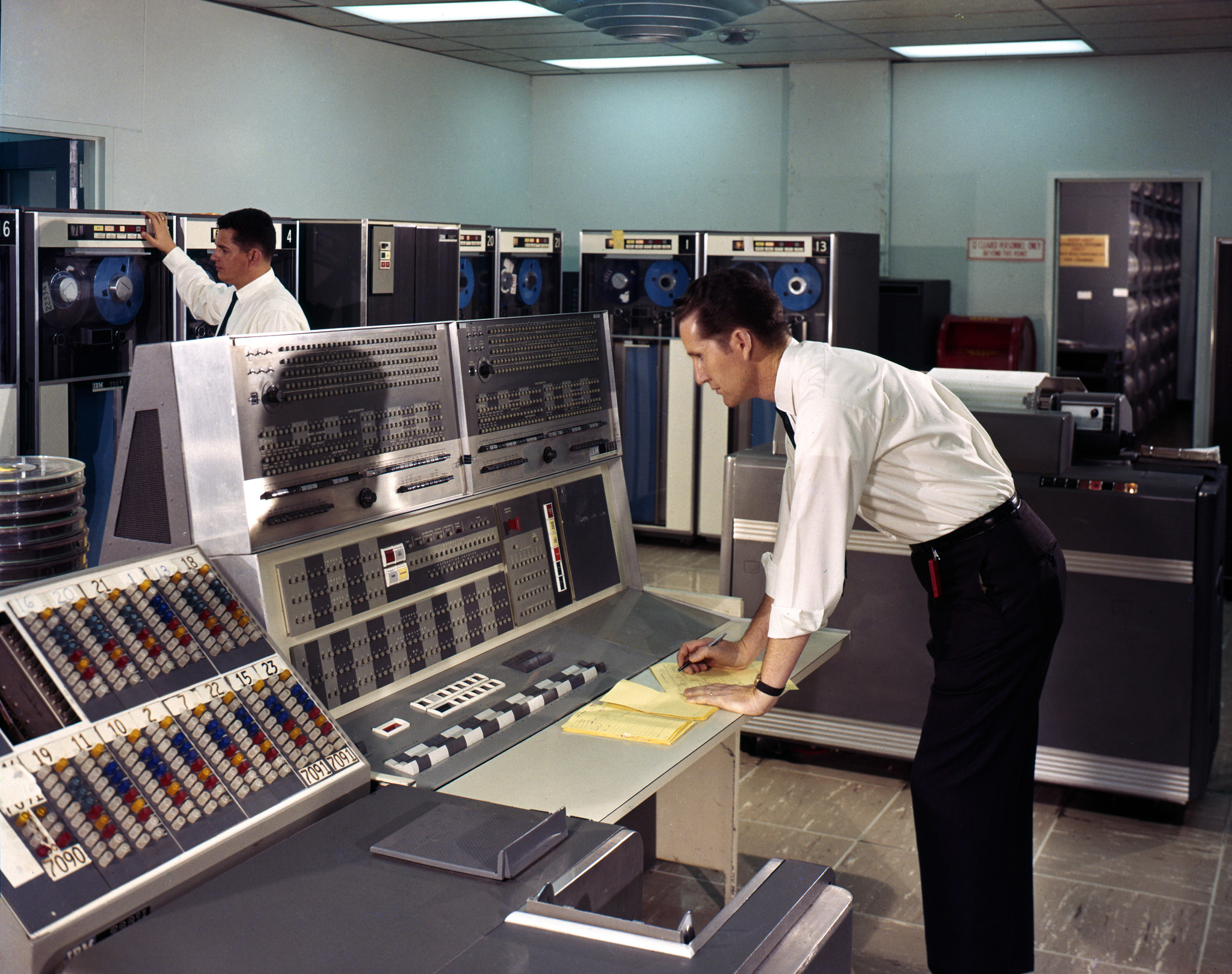 Two men in white dress shirts operate an IBM 7090 mainframe computer system in a large data centre. One man leans over the central operator's console writing on yellow notepad paper, while the other stands at a row of magnetic tape drive units in the background. The room is filled with large grey IBM mainframe cabinets and reel-to-reel tape drives. A numbered patch panel or channel selector is visible in the foreground left.