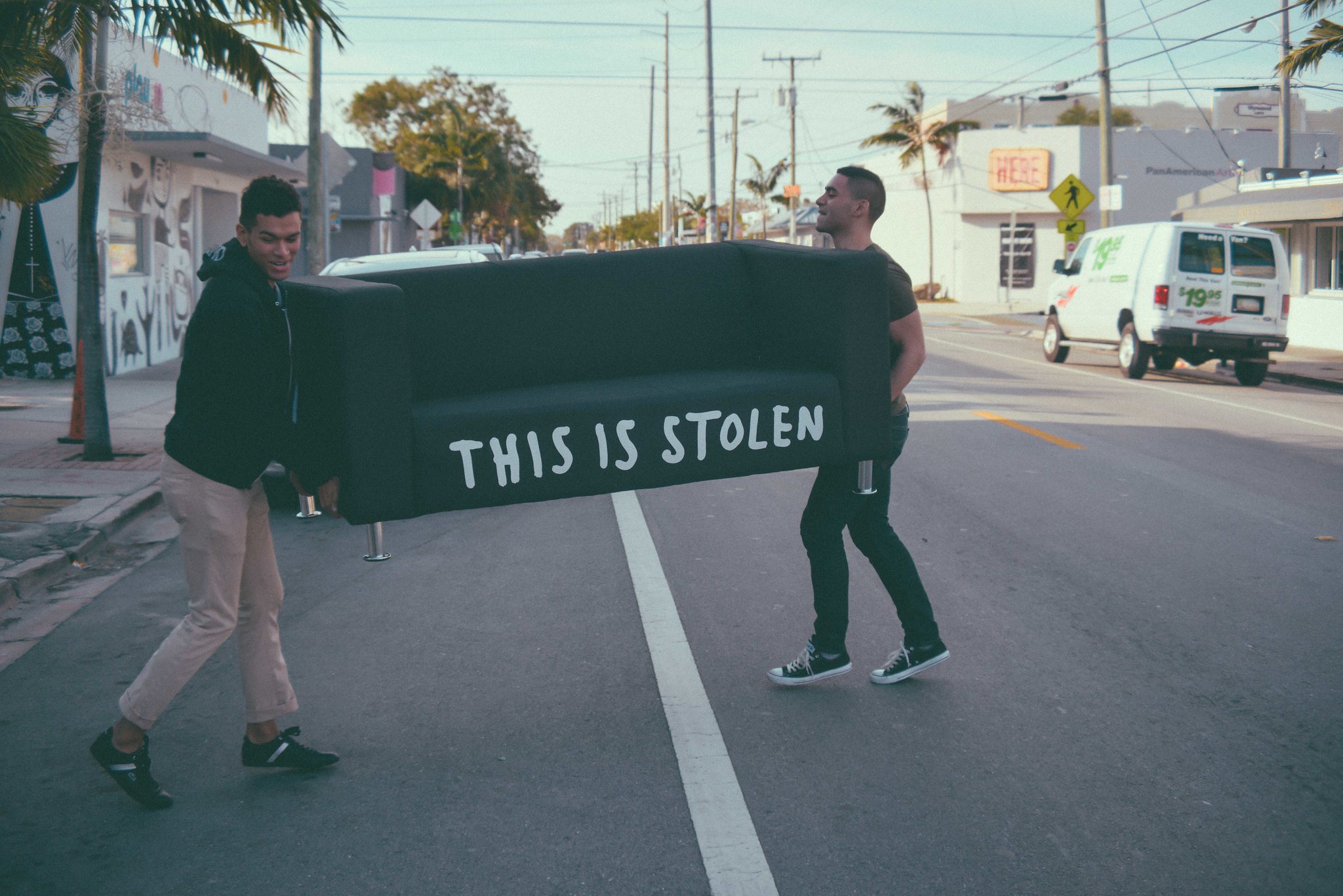 Two men carrying a dark green couch across a street in a tropical urban setting with palm trees. The couch has 'THIS IS STOLEN' written on it in large white hand-painted letters. Background shows commercial buildings, power lines, and vehicles on what appears to be a sunny day.