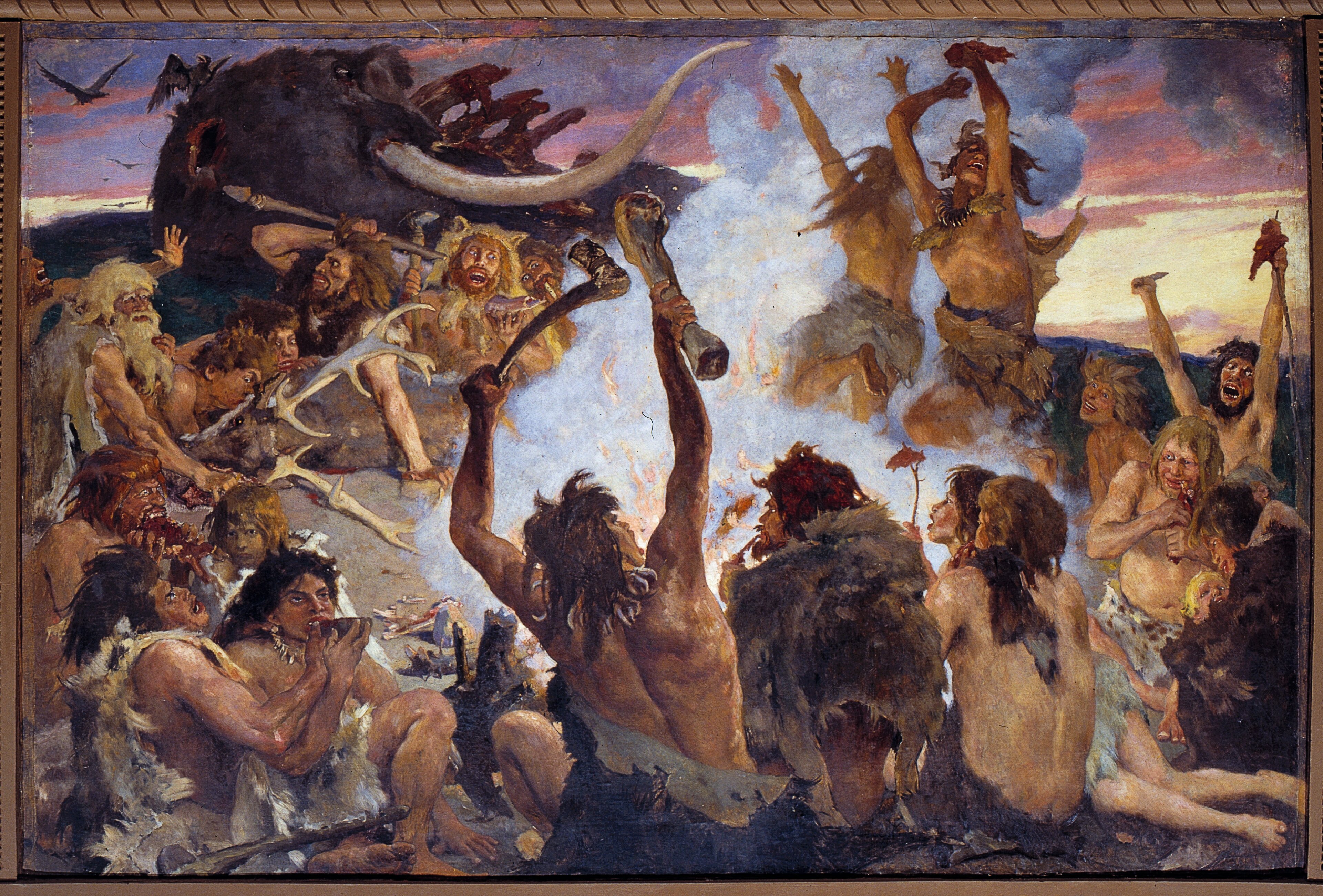 A dramatic 19th-century painting depicting a Stone Age scene with prehistoric humans gathered around a massive woolly mammoth. The central figure stands with arms raised holding a large bone or tusk, while others surround them in animated poses. Some figures work with mammoth tusks and bones in the foreground, while others celebrate or dance in the background. The scene is set against a dusky sky with birds flying overhead, and the composition conveys a sense of triumph and communal activity following a successful hunt.