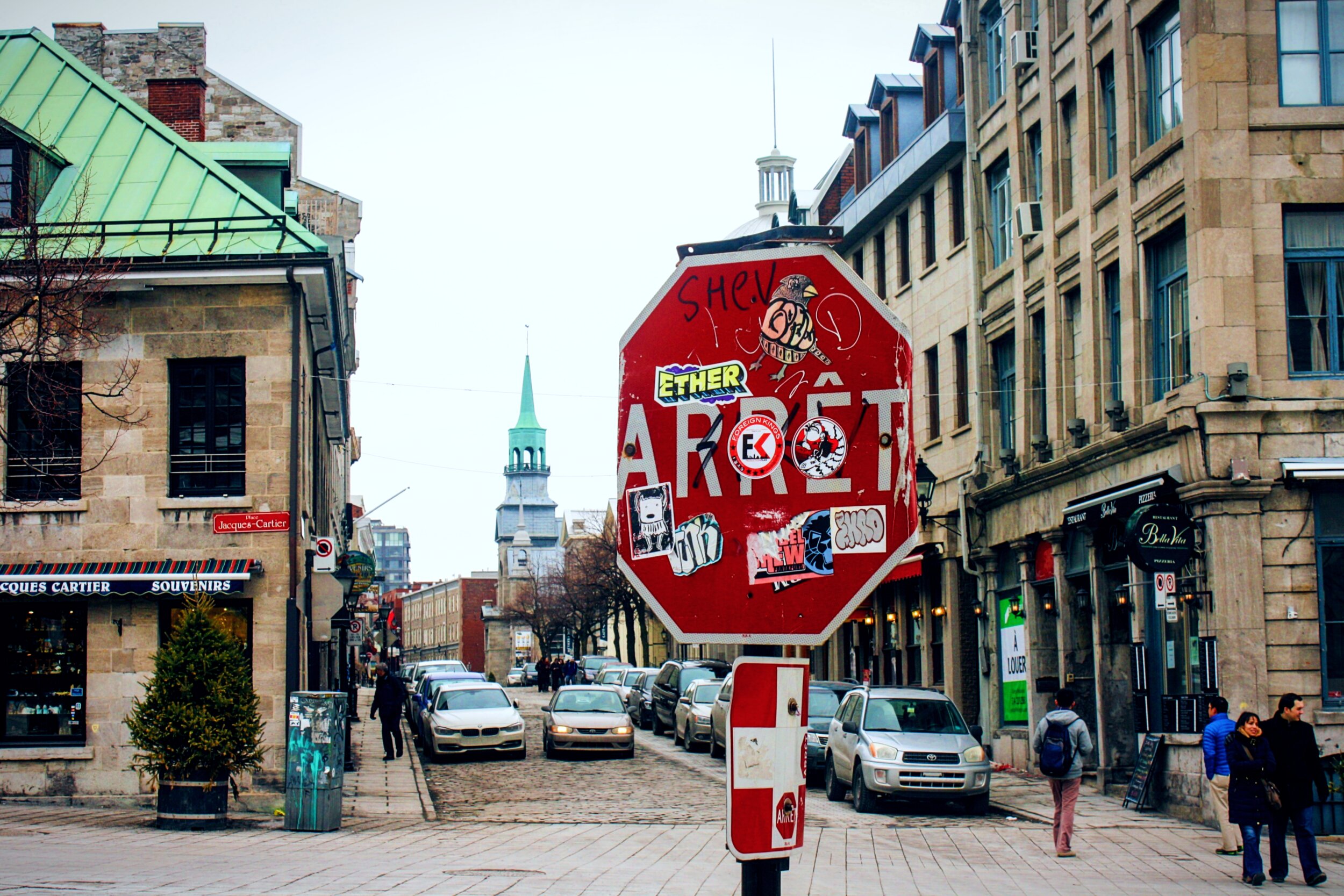 Un panneau d'arrêt rouge couvert de collants est planté au premier plan d'une rue pavée sur la place Jacques-Cartier, dans le Vieux-Montréal. Des deux bords, y'a des vieux bâtiments en pierre avec des toits mansardés pis des lucarnes, pis on aperçoit un clocher d'église au dôme vert en arrière-plan. La rue est pleine de chars stationnés pis d'autres en circulation, pis y'a quelques piétons qui marchent sur les trottoirs, près de boutiques comme la pizzeria « Bella Vita » pis le magasin « Jacques Cartier Souvenirs ». Le ciel est tout blanc et couvert, pis y'a un petit sapin en pot près de la devanture de gauche.