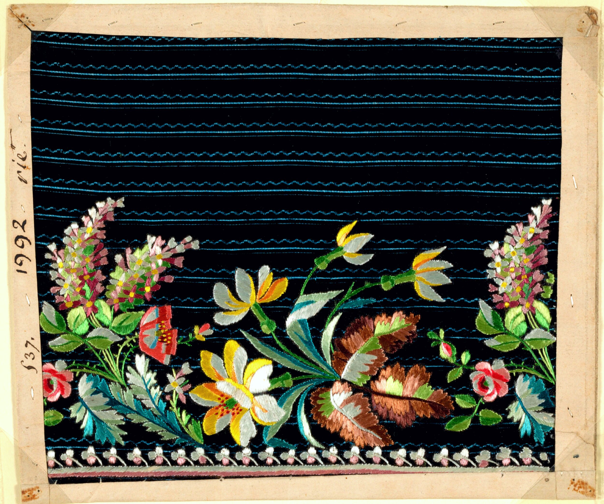 Antique textile sample mounted on a tan card, featuring black silk fabric with horizontal teal wave-stitched lines. Along the lower edge runs a colorful floral embroidery border with pink and yellow lilies, red poppies, pink roses, brown carnation-like blooms, and clusters of small pink and white flowers, connected by green and teal leaves. A row of small white and pink flowers forms a trim along the very bottom edge. Handwritten catalog markings '1992,' 'vie.,' and '537.' appear in the left margin.