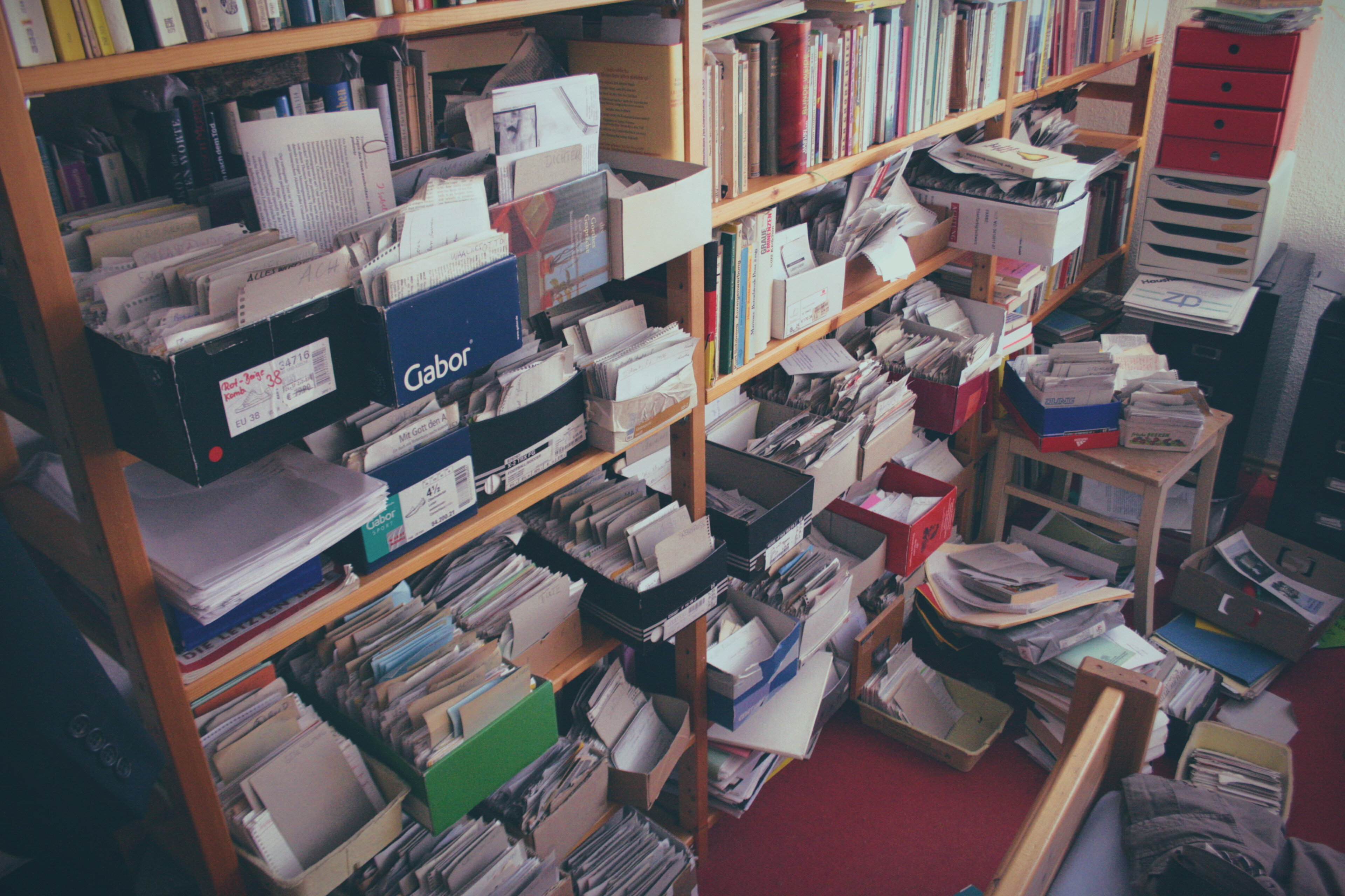 Wooden bookshelves overflowing with loose papers, documents, folders, and cardboard boxes including a Gabor shoe box. Additional stacks of papers and boxes are piled on a small side table and the red-carpeted floor; a knowledgeable home office or archive space.