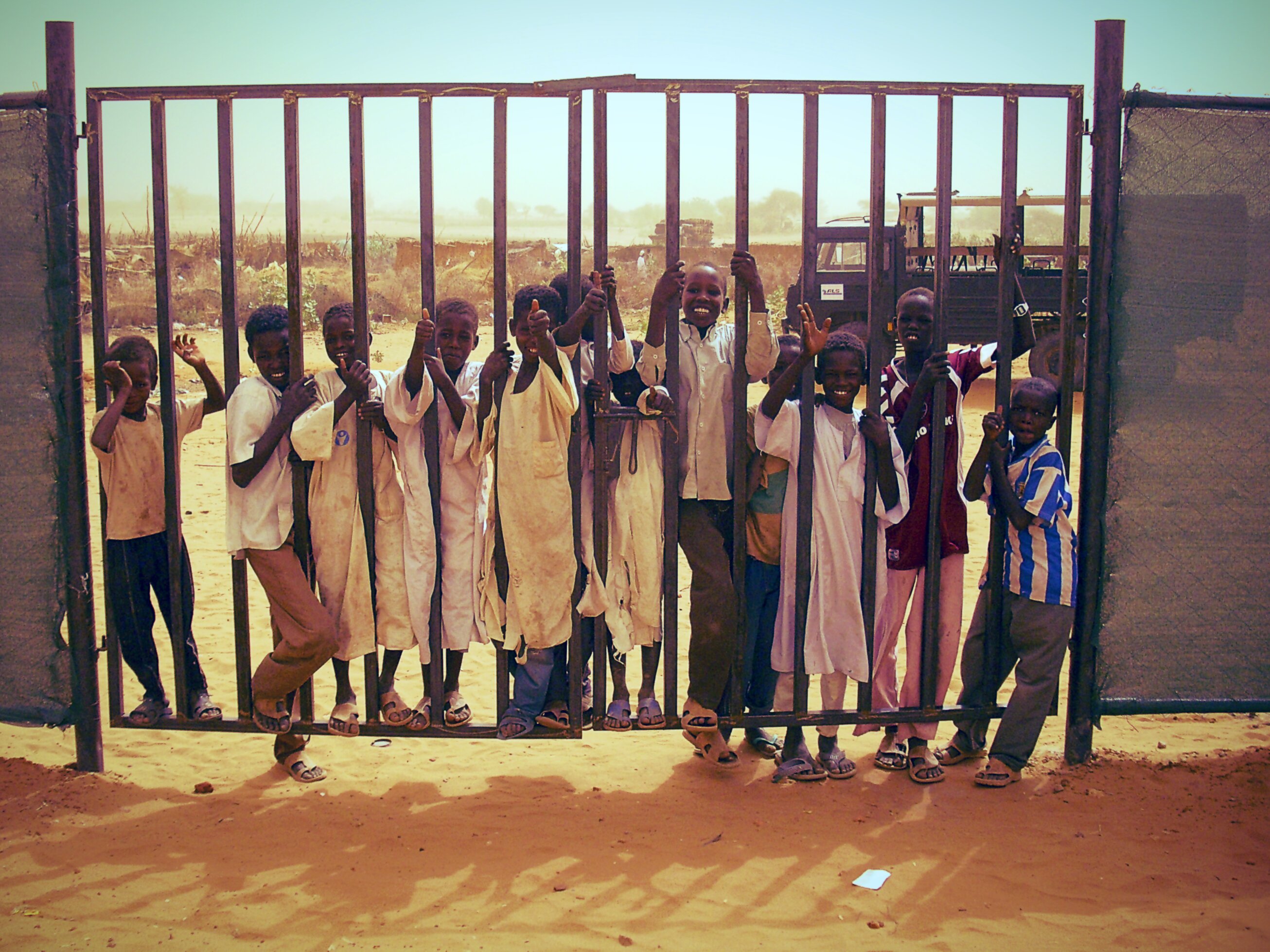 Twelve children of varying ages stand at and behind a large iron gate set in sandy ground. Most are smiling broadly and raising their hands toward the camera. Several wear white traditional robes and sandals. The background shows a flat, dusty landscape under a hazy blue sky