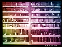 An archival photograph of a warehouse shelving unit divided into rows of numbered bins (125 through 739), each holding stacked metal bearings, bushings, and cylindrical machine parts. Many items have small paper tags with handwritten part numbers. The photo's colour has been altered into a diagonal rainbow cast, with magenta and pink across the upper left, shifting to blue in the upper right, and fading to yellow-green at the lower left. A partially legible cursive signature or stamp appears in the bottom right corner.