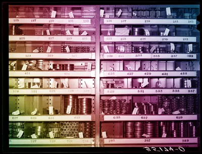 An archival photograph of a warehouse shelving unit divided into rows of numbered bins (125 through 739), each holding stacked metal bearings, bushings, and cylindrical machine parts. Many items have small paper tags with handwritten part numbers. The photo's colour has been altered into a diagonal rainbow cast, with magenta and pink across the upper left, shifting to blue in the upper right, and fading to yellow-green at the lower left. A partially legible cursive signature or stamp appears in the bottom right corner.