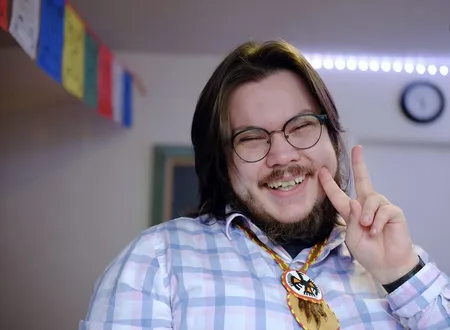 A smiling man (Brennan) with glasses, a mustache, and a beard, wearing a light plaid button-up shirt and a beaded pendant necklace with a thunderbird design, holds up a peace sign near his face. Colorful Buddhist prayer flags hang in the upper left, and a round clock is visible on the wall behind him.