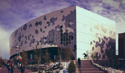 The Calgary Central Library, a striking curved building with a geometric glass and aluminum façade, on a partly cloudy autumn day. A skateboarder performs a trick on the front steps while pedestrians walk along the sidewalk below. Patches of snow are visible on the ground, and a colourful sculpture stands to the left of the entrance.