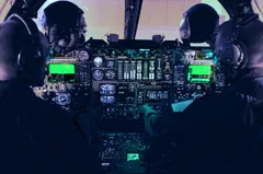 Two aircrew members wearing helmets, headsets, and oxygen masks are seated in a dimly lit military aircraft cockpit. The instrument panel between them glows with green multi-function displays, rows of analog engine gauges, and backlit switches, while light is visible through the windscreen ahead.