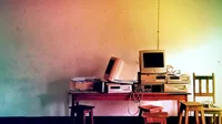A cluttered wooden table holds a row of obsolete beige desktop computers and CRT monitors, including a Dell and Compaq, with tangled cables, stacked against a stained wall lit in a pink-to-yellow-to-green gradient. Wooden stools and a chair sit in front of the table, suggesting an abandoned classroom or office.