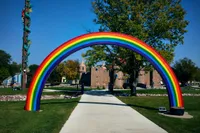 A large sculptural rainbow arch spans a concrete pathway in a park, painted in bright red, orange, yellow, green, blue, and purple stripes. In the background, a sandstone-colored castle-like building with turrets sits beyond a stone wall and small bridge, surrounded by green trees under a clear blue sky. To the left, a wooden pole wrapped in artificial vines features a small climbing figure, evoking a Jack and the Beanstalk theme.