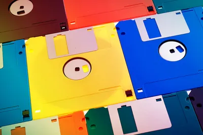 A collection of 3.5-inch floppy disks arranged in an overlapping, flat-lay pattern filling the entire frame. The disks span a wide range of colours — yellow, blue, red, teal, orange, dark brown, purple, and grey — creating a vibrant, saturated composition. The metal write-protect shutters and circular hub windows are visible on several disks, shot in close-up with bright, even lighting.
