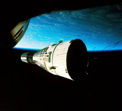 A spacecraft photographed from a companion capsule during orbital rendezvous, its white and black cylindrical body lit against the blue curve of Earth's atmosphere. The photographing craft's window frame cuts across the upper left corner, and thin antenna wires trail from the target vehicle's rear section into the black of space.