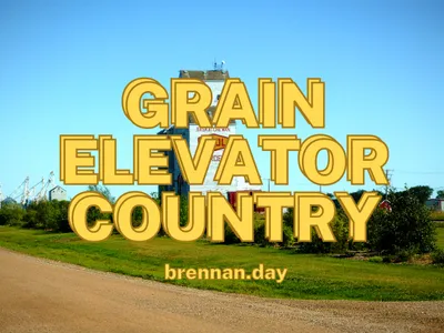 Bold yellow outlined text reading 'GRAIN ELEVATOR COUNTRY' in large stacked letters, with 'brennan.day' in smaller bold yellow text below. The background photograph shows a white wooden grain elevator marked 'SASKATCHEWAN' in a small prairie town, flanked by green trees and shrubs under a clear blue sky. A curved gravel road runs along the bottom foreground, with additional grain storage structures visible to the left and power lines to the right.