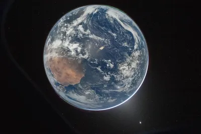 A photograph of Earth from deep space, slightly off-centre and partially in shadow, with a thin luminous blue atmospheric halo visible along the sunlit edge. Australia's arid interior is visible as a rust-coloured landmass on the left side. Swirling cloud systems cover much of the Pacific Ocean to the right. Stars are faintly visible in the black background, and a bright lens flare appears near the lower right.