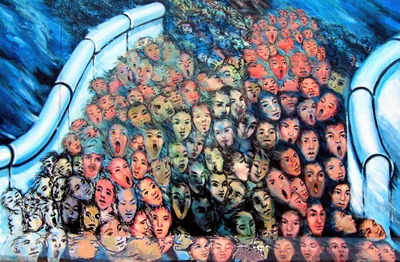 Mural of a dense crowd of painted faces surging like a wave through a gap in pale blue, pipe-like walls, set against a deep blue background. Some faces have closed eyes or calm, wary expressions, while others have wide-open mouths, as if shouting or singing. Skin tones run from orange and pink to green, teal, and gray, and the faces along the bottom edge are reflected, as if in water.