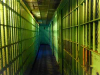 A narrow jail corridor washed in green light, with floor-to-ceiling barred cells on both sides and a slatted metal ceiling above. Orange rust shows through the worn paint on a few bars at lower right, and the hallway ends at a blank wall