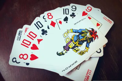 Several playing cards fanned out face-up on a dark wood surface. Visible cards from left to right include the 8 of Hearts, 10 of Hearts, 10 of Clubs, 8 of Diamonds, 10 of Spades, 9 of Clubs, and 9 of Hearts. Prominently displayed near the center is a Joker card depicting a jester figure in a colorful blue, green, and gold costume with a red and black jester's cap, holding letter blocks. Additional Joker and number cards are partially visible beneath the spread. All cards bear the text '117 Anos — Cuidando de Você,' a branded Brazilian playing card deck.
