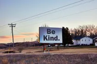 A large roadside billboard reading 'Be Kind.' in bold black letters on a white background, mounted on a 'Newman' sign structure in a grassy field at dusk. A wooden power pole with lines runs along the left, with rolling hills, a house, evergreen and bare trees, and a soft pink-and-blue sunset sky in the background.