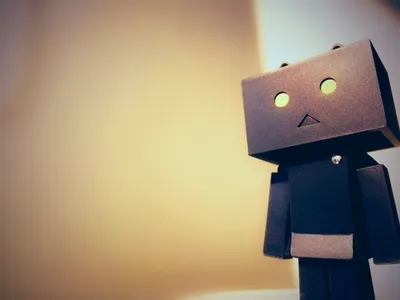 A cardboard box robot figure (Danbo-style) with round glowing yellow eyes and a triangular mouth, standing against a warm, softly blurred beige and cream background. The figure is positioned on the right side of the frame, gazing off to the left, with the left portion of the image left empty and softly lit.