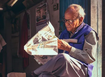 An elderly man sits in morning sunlight outside a weathered blue doorway in Basantapur, Nepal, absorbed in reading a Nepali-language newspaper. He wears black-rimmed glasses, a blue vest over a checked shirt, and light grey trousers. The newspaper catches the early light brilliantly against the dark background. A cane rests under his arm.