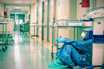 A healthcare worker in full protective equipment, including a blue gown, surgical cap, face mask, and gloves, sits exhausted on the floor of a hospital hallway, leaning against a medical cart. The corridor stretches behind them with empty gurneys and medical equipment visible along the pale walls under fluorescent lighting, conveying the physical and emotional toll of frontline medical work.