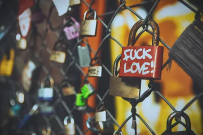Close-up photograph of numerous padlocks attached to a chain-link fence, with a red lock in sharp focus bearing the handwritten message 'FUCK LOVE!' in white paint. The surrounding locks are blurred by a shallow depth of field, while warm orange and yellow tones in the background create a soft, out-of-focus glow. The featured lock shows signs of rust and wear, contrasting with the romantic 'love lock' tradition typically associated with such fences.