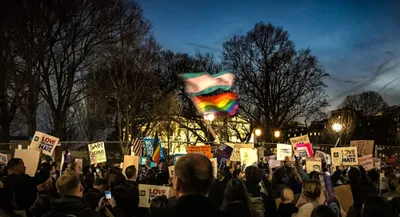 A crowd of protesters gathered at dusk holding signs and waving transgender pride and rainbow flags. Visible signs read 'LOVE TRUMPS HATE,' 'TRANS RIGHTS,' 'SAD!,' and 'NO HATE.' Bare trees and illuminated street lights frame the scene against a blue evening sky, with buildings visible in the background.